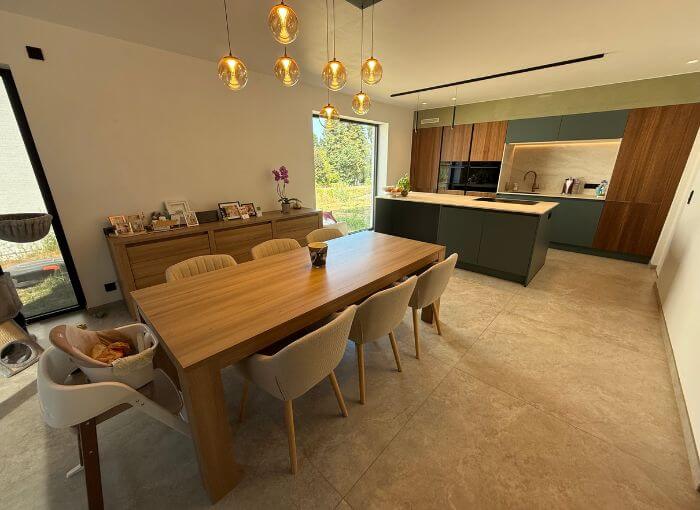 Cuisine et coin repas modernes avec une table en bois, six chaises, des suspensions, un îlot de cuisine, des placards, une chaise haute, un buffet orné de photos et une fenêtre donnant sur la verdure à l'extérieur.