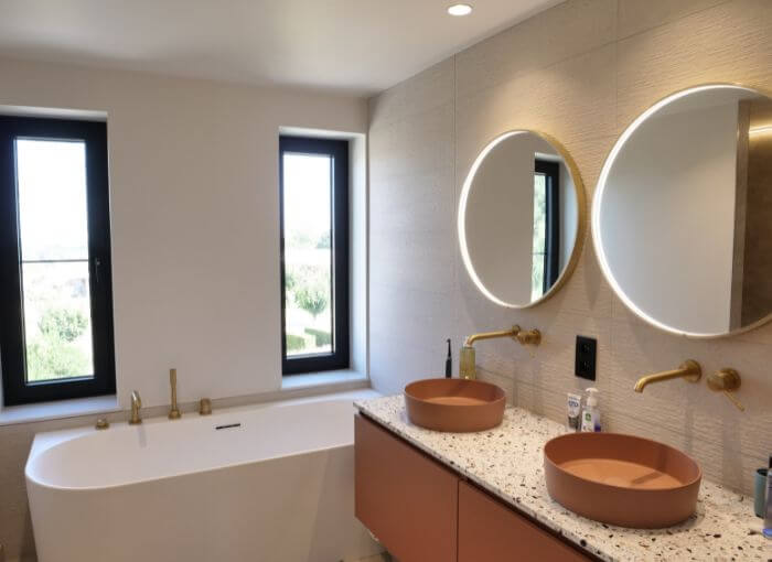 Salle de bains moderne avec une baignoire blanche placée sous deux fenêtres, une double vasque avec des miroirs ronds, des lavabos sur pied marron, des robinets dorés et un plan de travail en terrazzo.