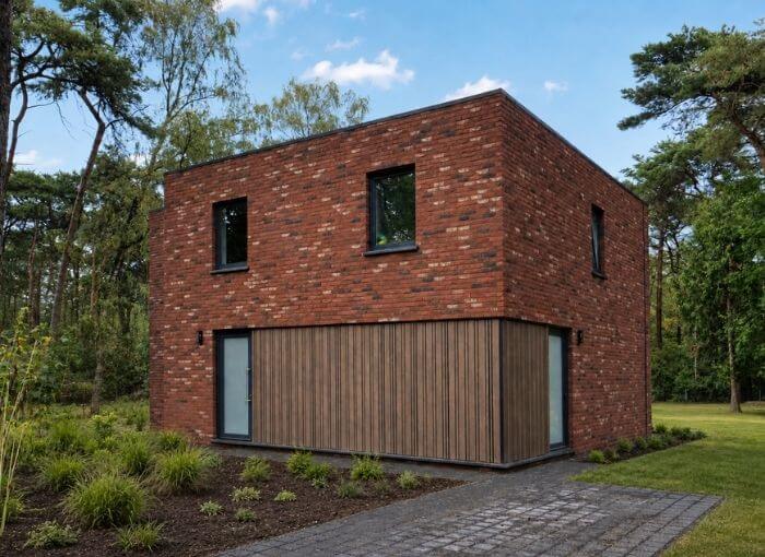 Une maison moderne à deux étages, aux murs de briques rouges et aux boiseries, entourée de verdure et d'arbres sous un ciel bleu.