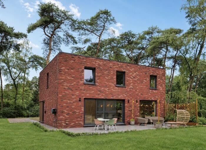 Une maison moderne en briques, à deux étages, dotée de grandes fenêtres, se dresse au milieu de grands arbres. Au premier plan, on aperçoit une terrasse équipée de mobilier d'extérieur et une pelouse verte.