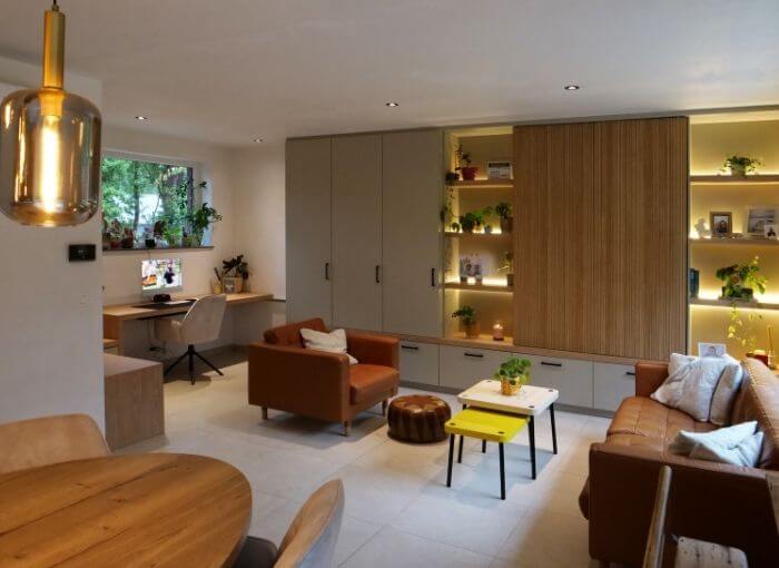 Un salon moderne avec des canapés beige clair, une table basse jaune, des plantes et des étagères encastrées éclairées par un éclairage chaleureux. À l'arrière-plan, on aperçoit un petit coin bureau avec un bureau et une fenêtre.