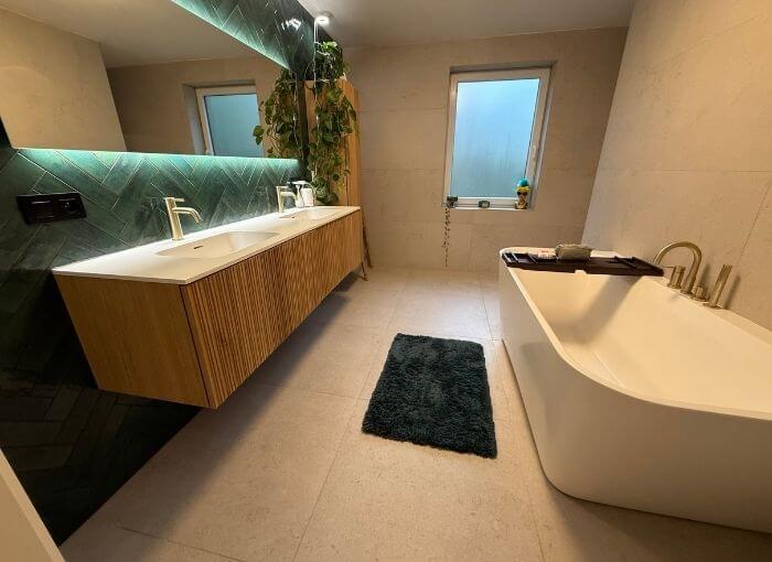 Salle de bains moderne avec un meuble-lavabo suspendu à double vasque, un grand mur de miroir carrelé de vert, une baignoire rectangulaire, un tapis foncé, une fenêtre dépolie et une grande plante dans un coin.